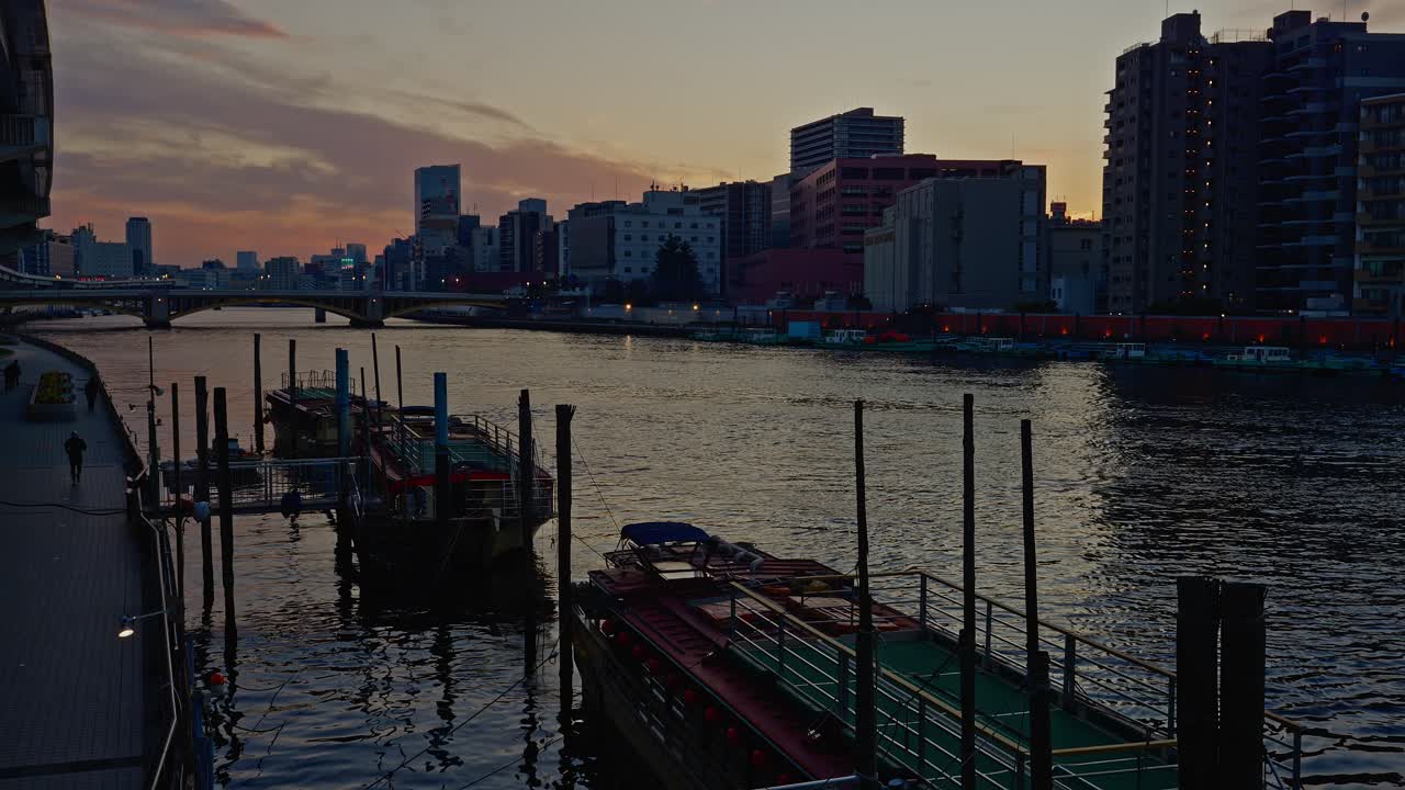 En lugn solnedgångsscen över Sumidafloden i Tokyo. Traditionella Yakatabune-husbåtar ligger förtöjda längs banken med en modern stadssiluett och skymningshimmel.