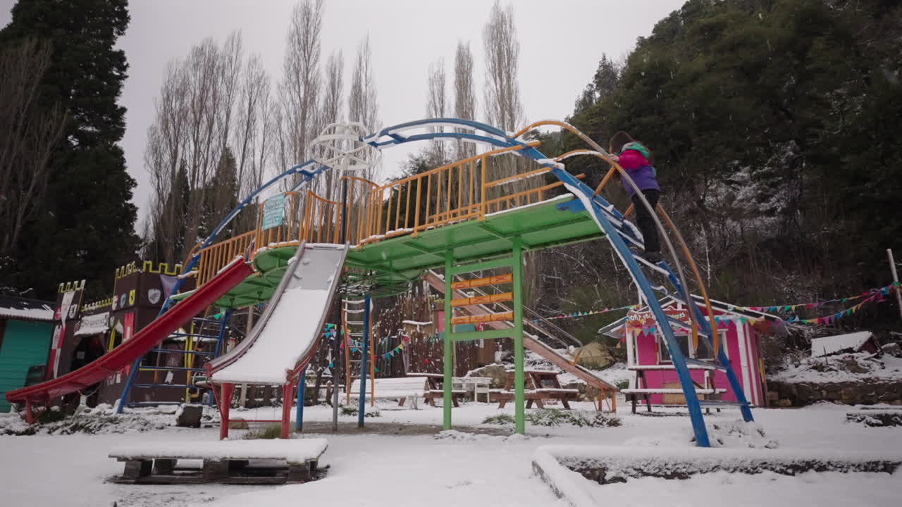 Static shot in Colonia Suiza, Bariloche, Argentina, showing a child climbing a snowy metal playground structure amid colorful play equipment and tall poplars after a recent winter snowfall.