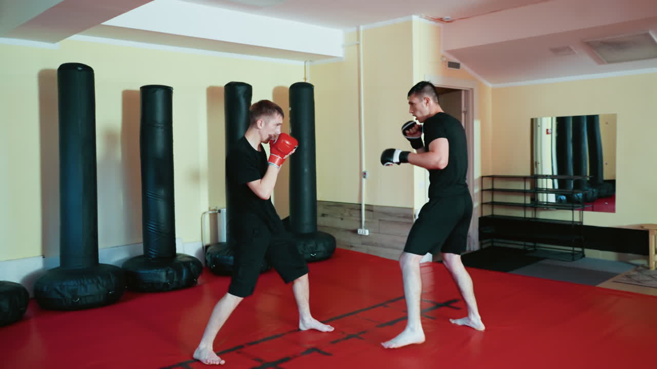 Wrestlers sparring inside gym, wearing gloves and black outfits, practicing combat sport stance and preparing for martial training session on red mat floor surrounded by standing punching bags
