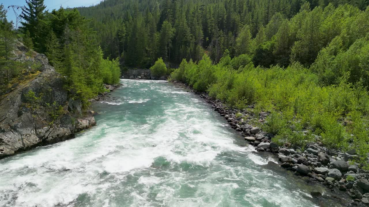 An aerial view of a fast-flowing river cutting through dense evergreen forest with rocky banks and distant mountains near Squamish, BC, Canada