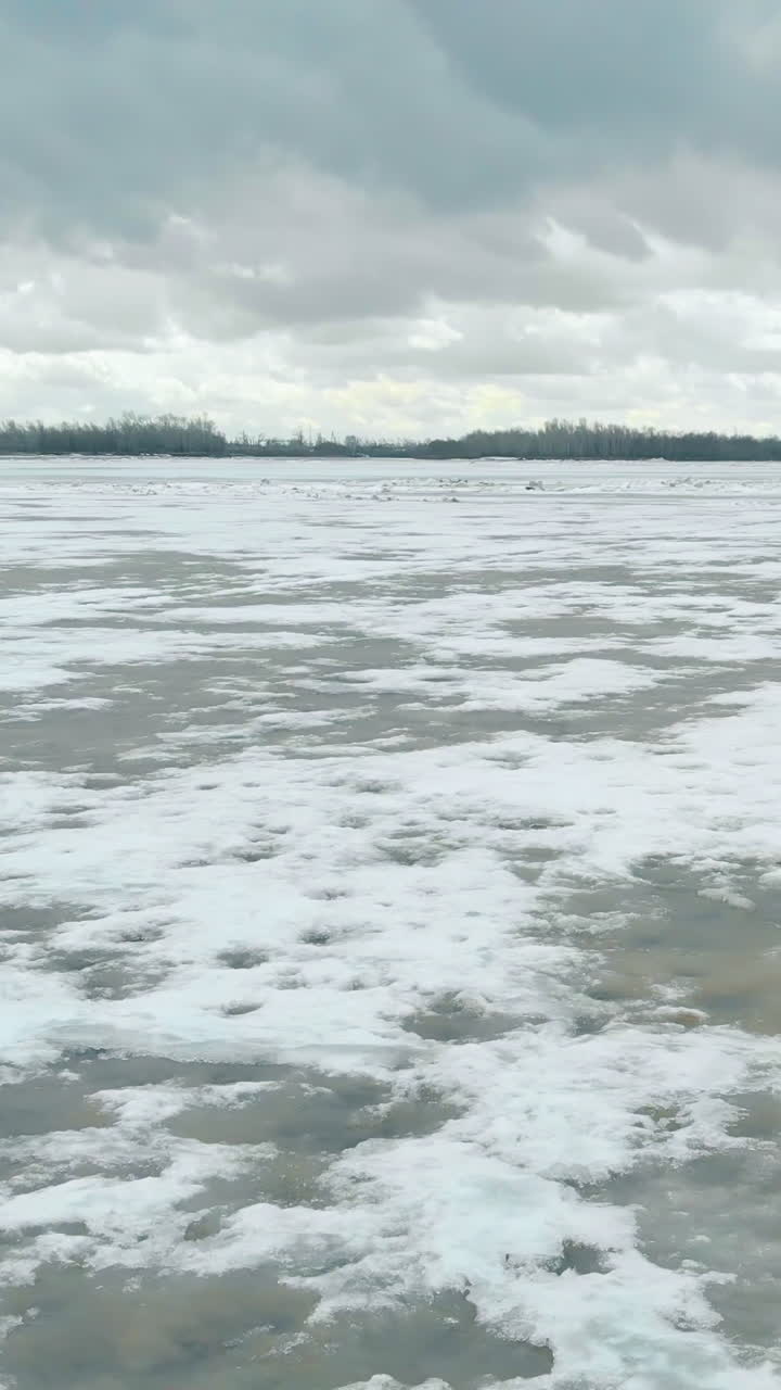 wide river with white ice surrounded by dense hilly forests under sky with grey clouds in spring upper view