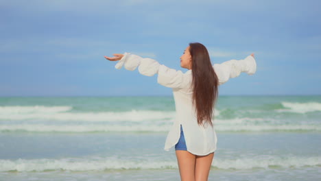 back view of brunette asian girl on seashore raising her arms to the sky