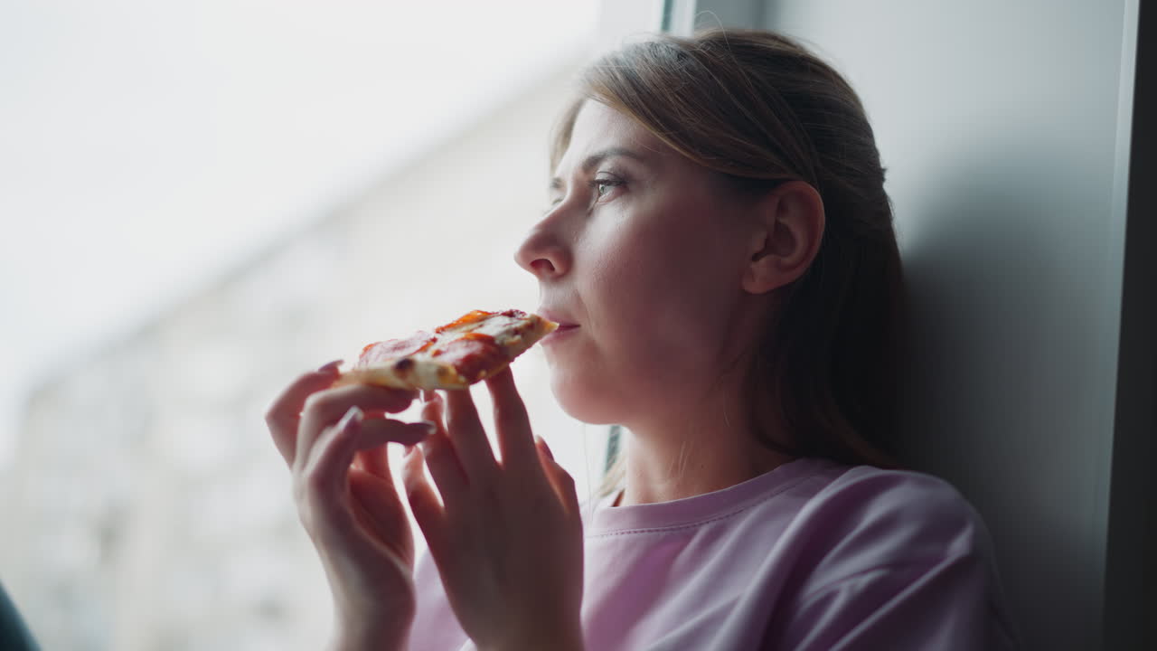Girl wearing purple shirt looking through glass window while holding slice of pizza, thoughtful expression on face, soft daylight filtering in from outside