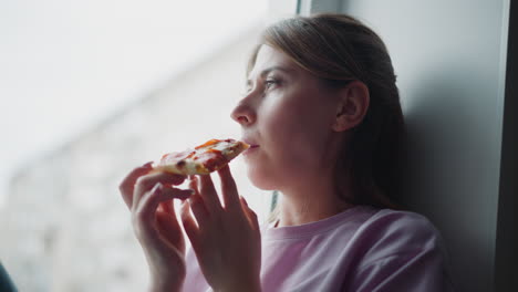 girl wearing purple shirt looking through glass window while holding slice of pizza, thoughtful expression on face, soft daylight filtering in from outside