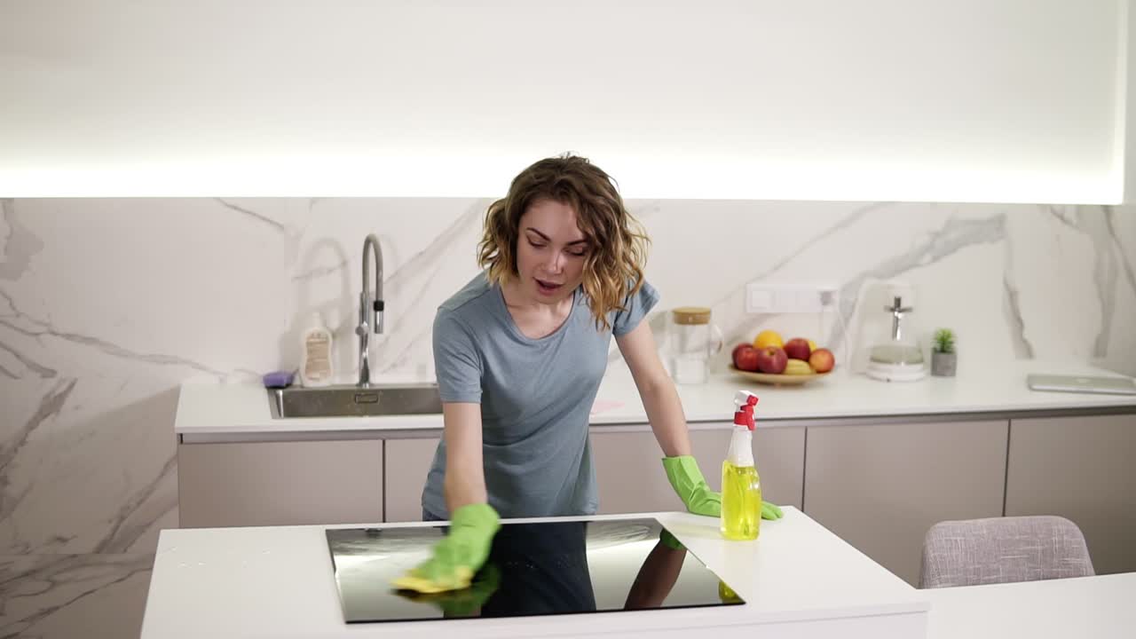 chica caucásica limpiando la estufa de cocina el panel de cocina en la cocina. mujer joven limpiando la superficie perfecta de la estufa de cocina de cerámica negra y el mostrador usando guantes y trapeador. divirtiéndose y cantando. cámara lenta