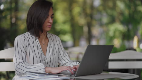 mujer de negocios morena grupo étnico hispano está escribiendo en una computadora portátil mientras está sentada en una mesa en una cafetería de verano haciendo trabajo remoto. mujer independiente. pequeña empresa