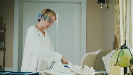 woman in a bathrobe with curlers on her head ironing clothes