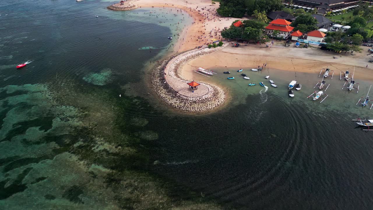 日中の熱帯ビーチを空撮ドローンで捉えた眺め。湾曲した石の防波堤が、浅い礁湖のあるビーチを保護しており、穏やかな水辺では人々が泳ぎ、砂浜の海岸線には小さなボートが停泊しています。