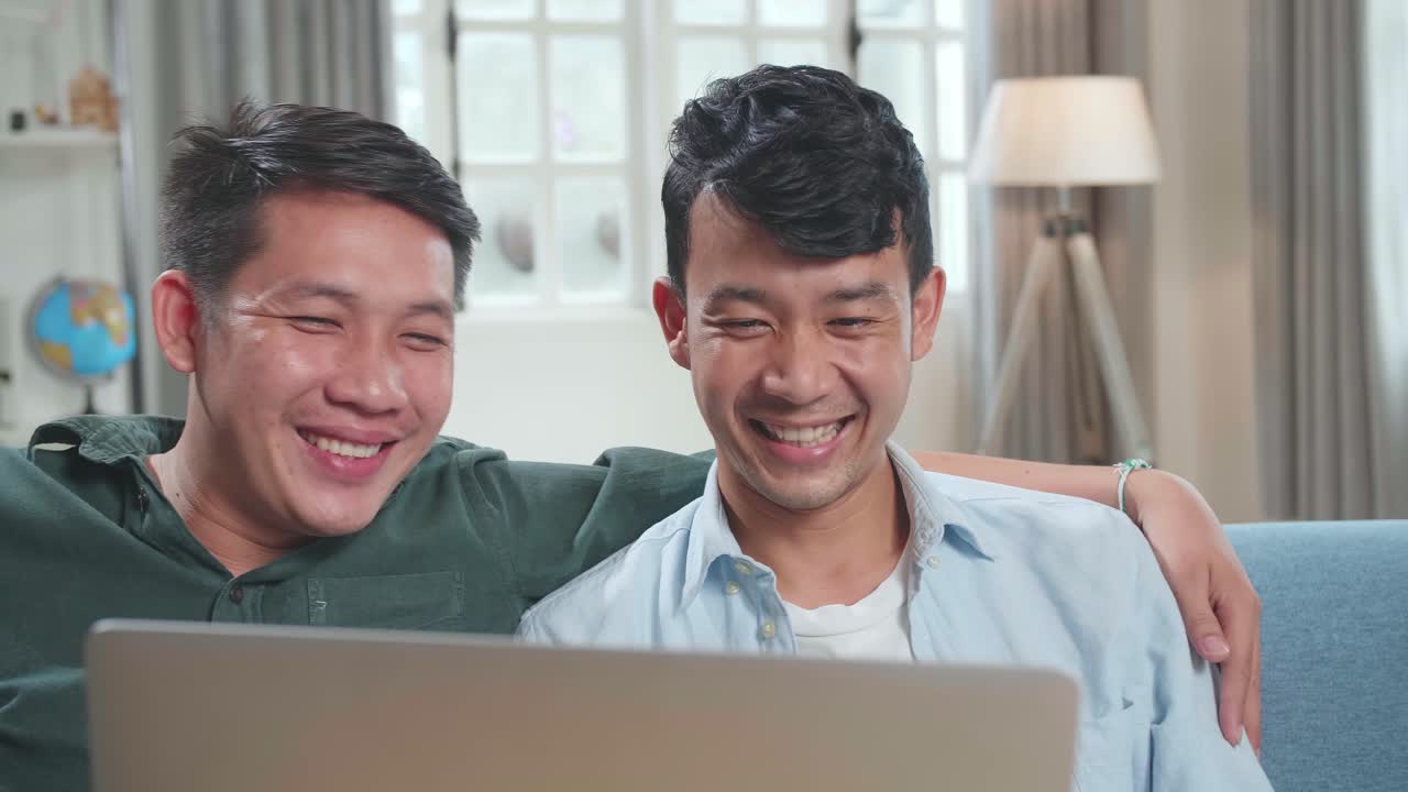 Portrait Of Asian Gentle Gay Couple Using Laptop Computer, While Sitting On A Couch In Cozy Stylish Apartment. Adult Boyfriends Online Shopping On Internet, Watching Funny Videos On Streaming Service