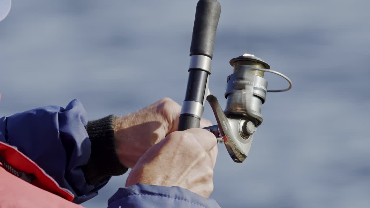 Close-up of an elderly fisherman's hands winding a reel, blurred in background