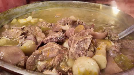 close-up shot of a metal pot cooking sancocho, the traditional dominican dish. a metal spoon stirs the mix, highlighting the meats and vegetables as steam rises.