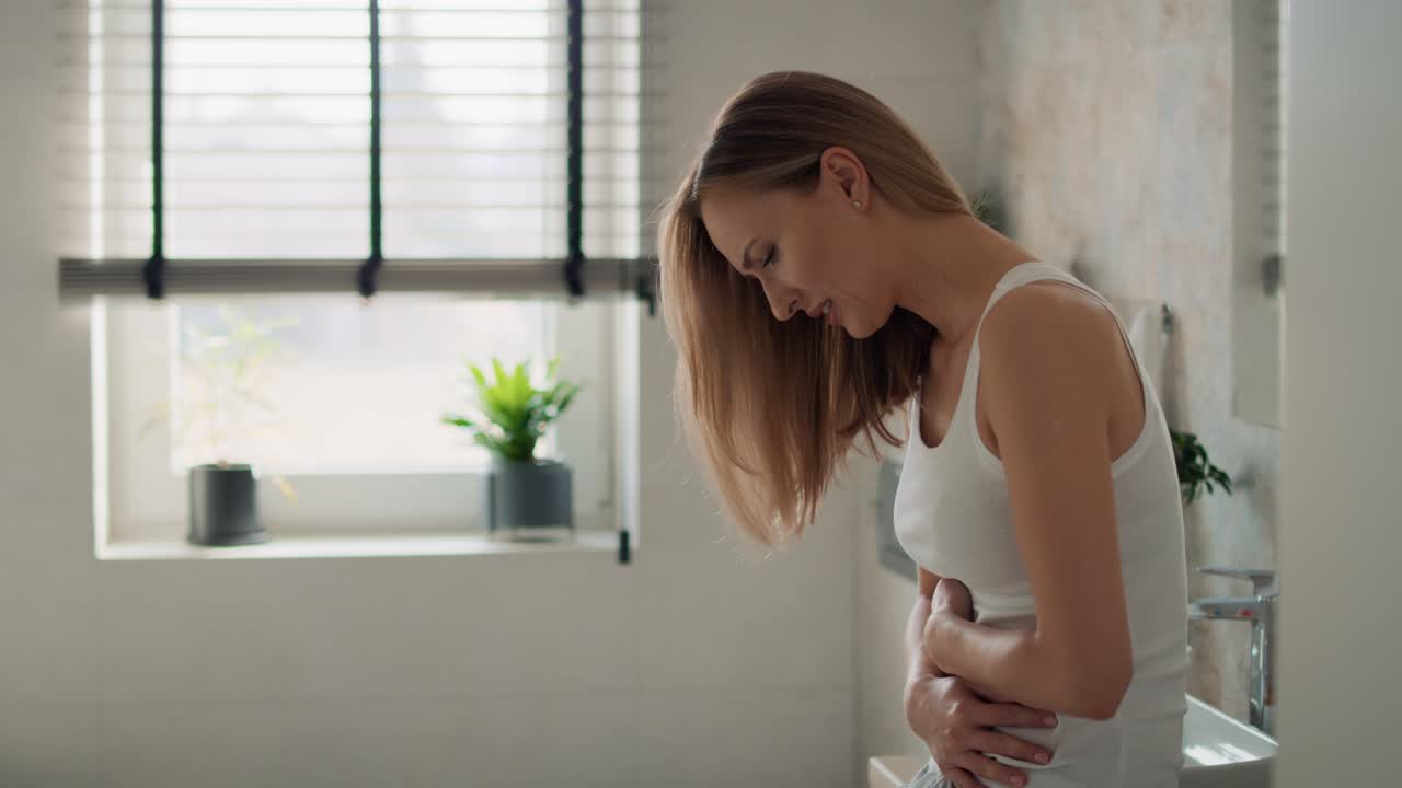 Young caucasian woman standing in the bathroom and feeling pain in the stomach.