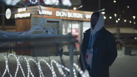 person standing in front of glowing white christmas lights in urban setting at night. shop signage visible in background with lighted decorations. winter evening scene with festive atmosphere