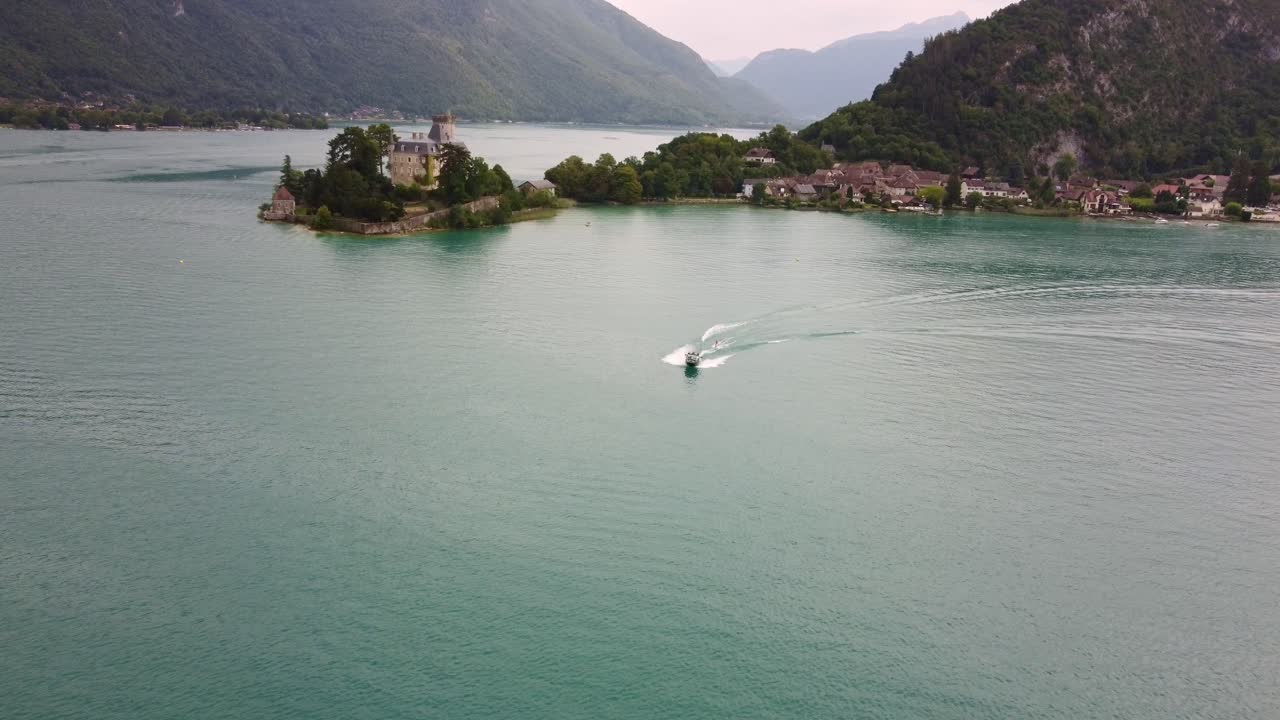 A speed boat towing a wake boarder next to Château de Duingt on Lake Annecy. Amazing scene with the boat shooting across the lake with amazing views in the background. Boat is heading towards