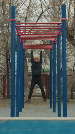 a coach swings on a red iron bar in an outdoor fitness area, with people playing football in the background, surrounded by bare trees and residential buildings