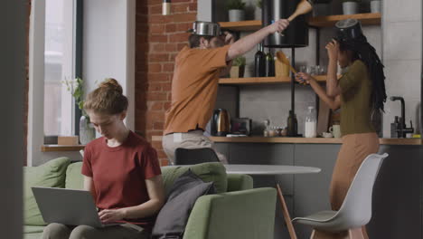 girl working on laptop computer sitting on sofa while her two roommates having fun fighting with kitchen utensils