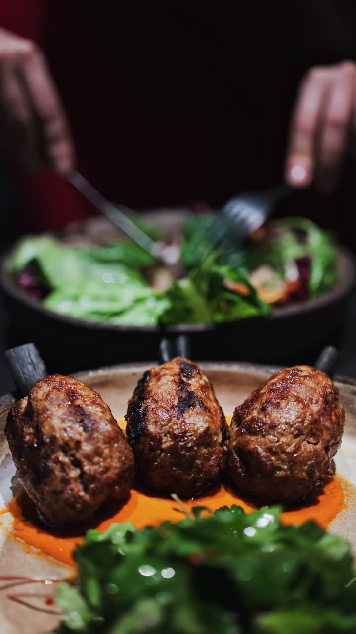 Three grilled meat skewers on charcoal sticks served on a plate with an orange sauce with a woman cutting and eating a salad at a restaurant on the background