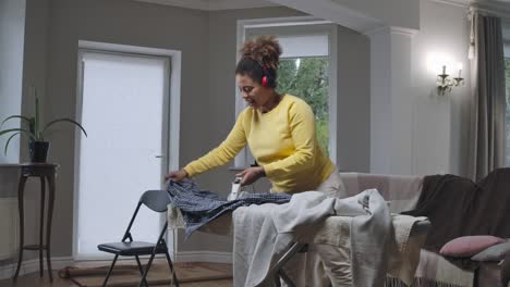 wide shot of joyful african american woman ironing laundry dancing and singing to music in headphones. happy cheerful relaxed housewife enjoying housekeeping. cinema 4k prores hq.