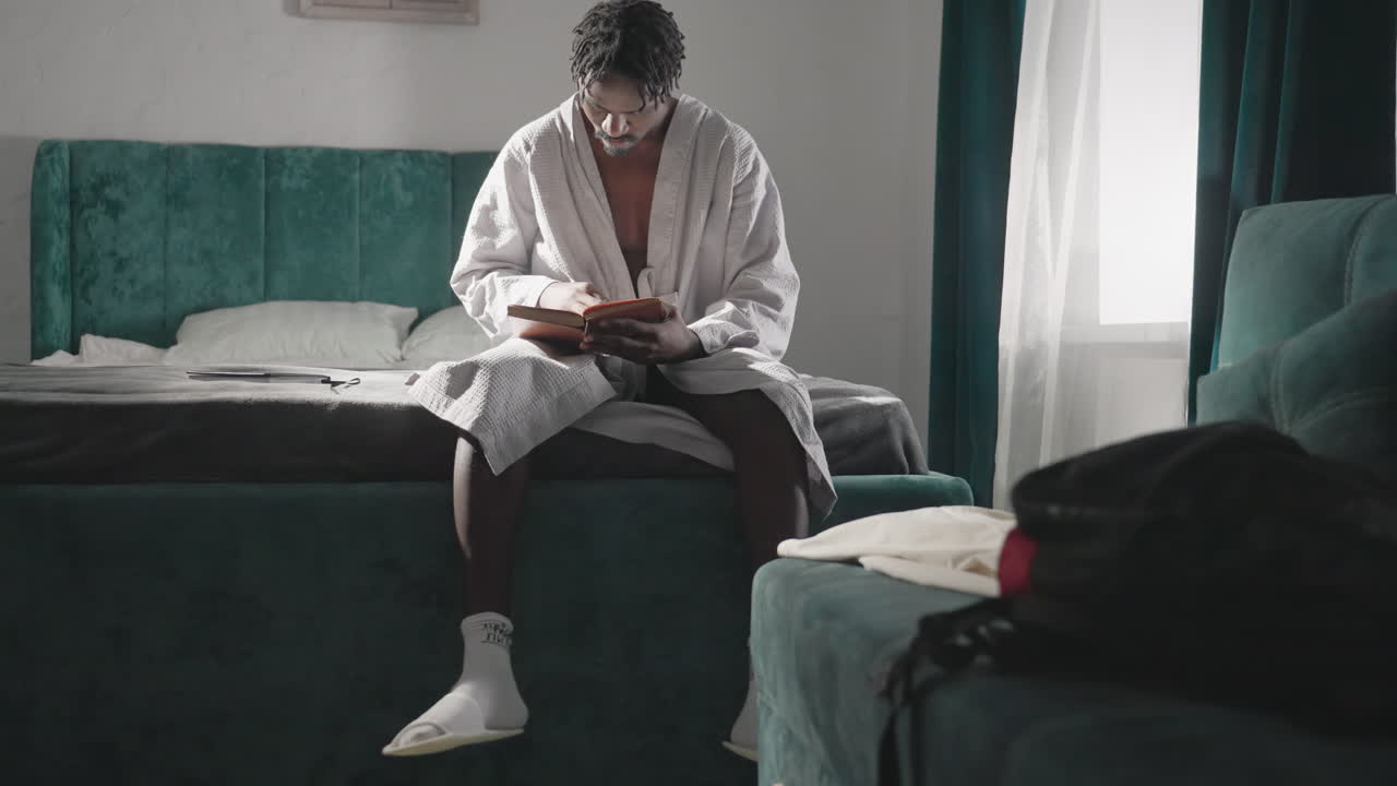 Man in white robe sitting on bed, deeply focused on reading book, turning pages slowly. Soft natural light from window adds to calm, peaceful atmosphere in serene indoor setting