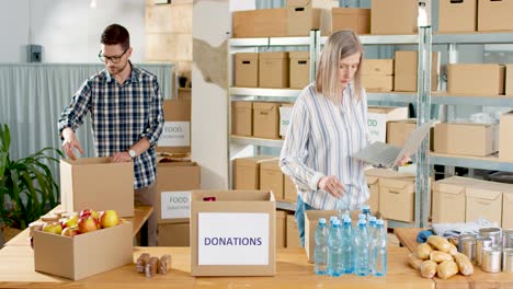 Young-caucasian-male-volunteer-putting-food-in-a-donation-box-in-charity-warehouse-while-senior-woman-typing-on-laptop