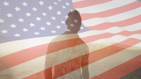 animation of american flag waving over african american man standing on beach