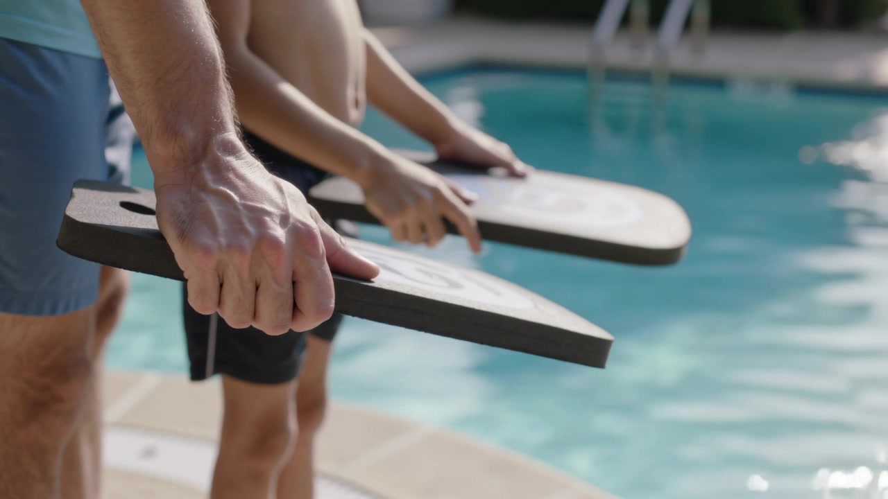 Holding kickboards, three shirtless men in swim trunks leaning toward pool edge, ladder visible