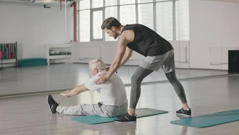 Fitness-coach-stretching-to-elderly-man-in-sport-club.
