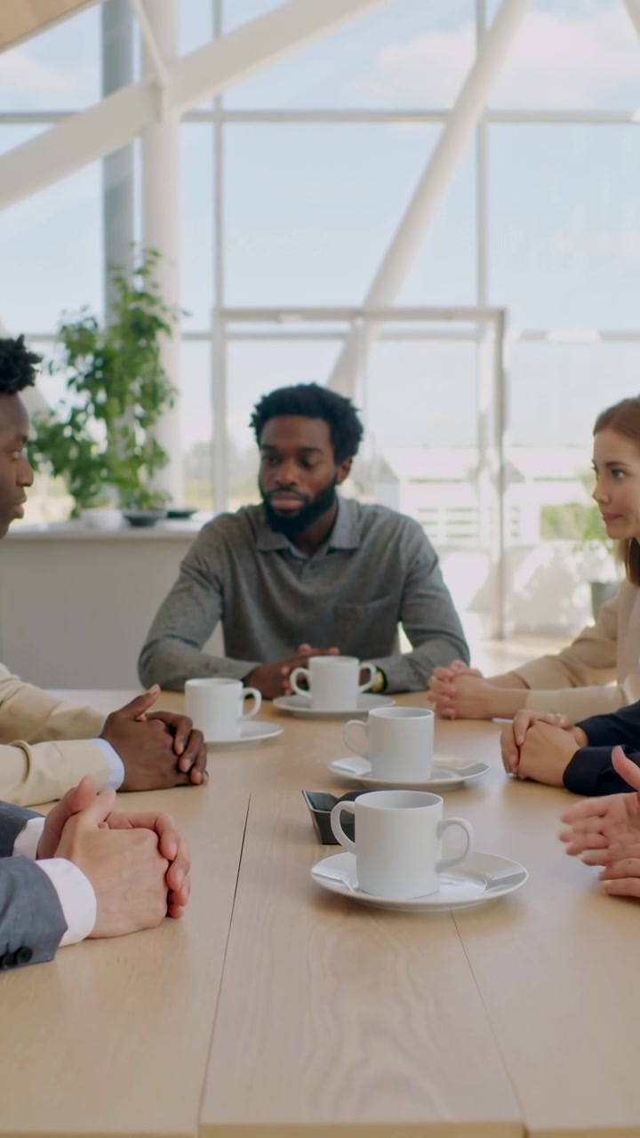 Verticale video: Luisterende man in grijze polo bij kopje, terwijl collega rechts begint te praten op kantoor tijdens koffie.
