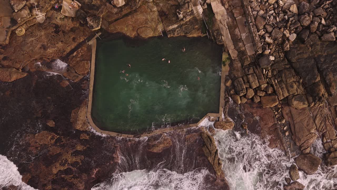 Aerial top-down view of swimmers in ocean rock pool with winter waves crashing around – dramatic coastal scenery and seasonal contrast in nature and lifestyle.