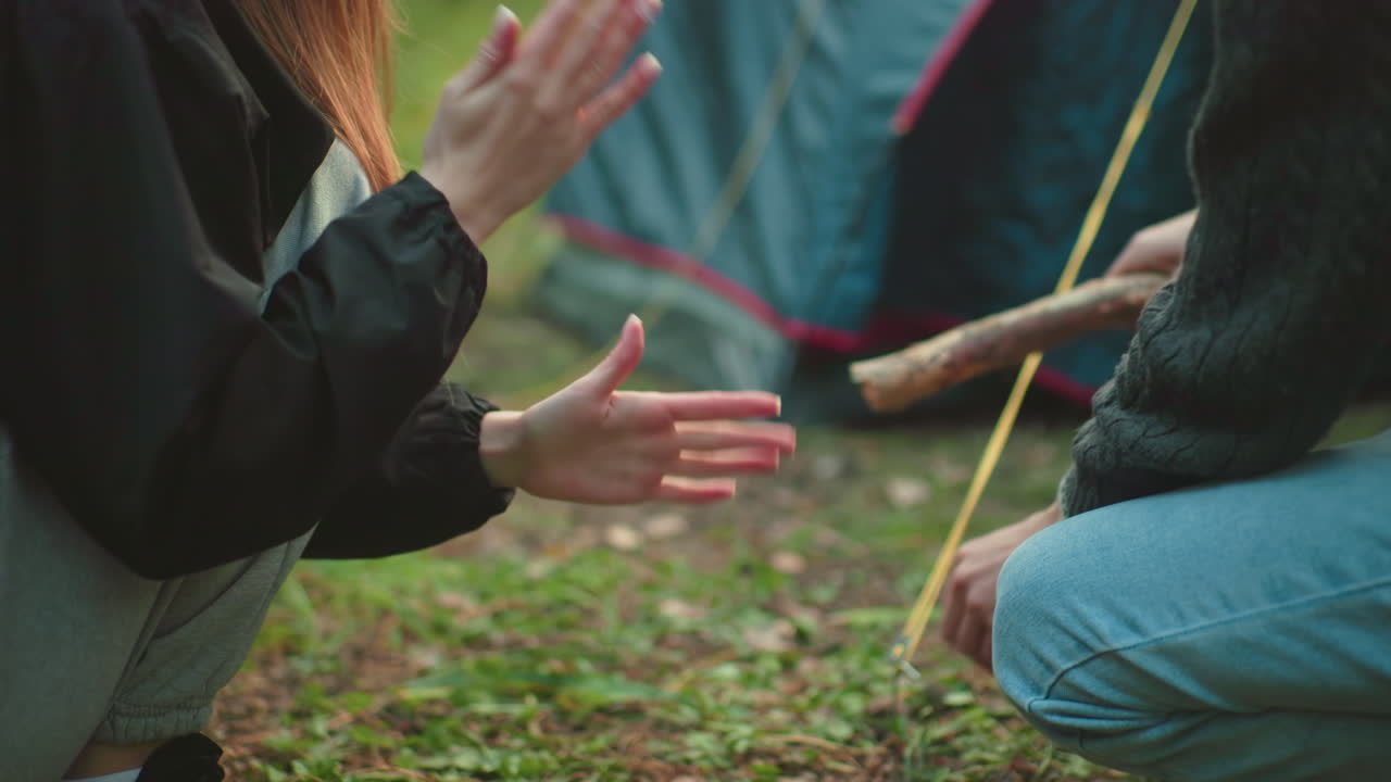 Close up of woman dusting hands as man beside her uses wooden stick to hammer tent peg into grassy ground with ropes extending from nearby tent, showcasing teamwork