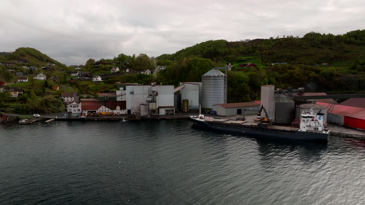 Freight boat delivers soy cargo near industrial shoreline with green hills, aerial establishing over water
