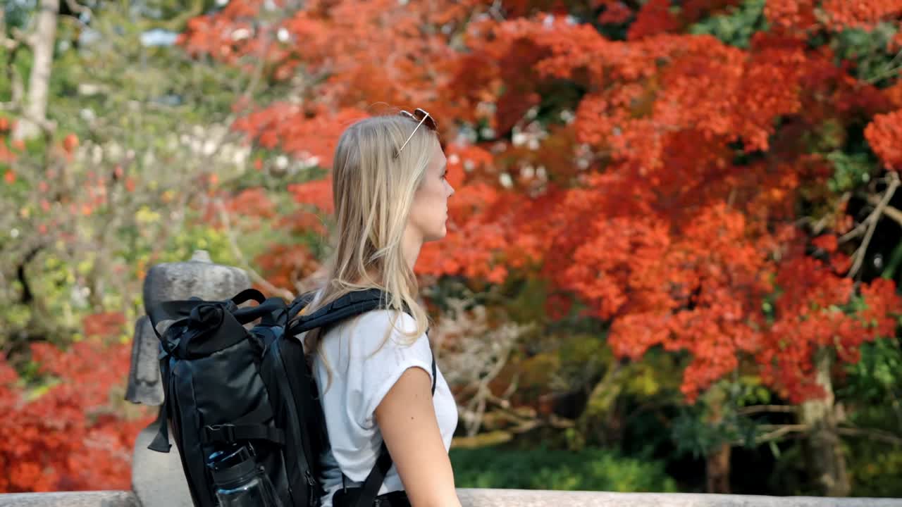 A young woman walks across a traditional wooden bridge in Kyoto, Japan, surrounded by vibrant red autumn foliage.