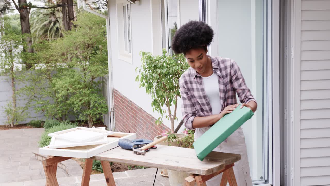 African American woman reaching frames, lifting, prepping green frame at porch bench, copy space