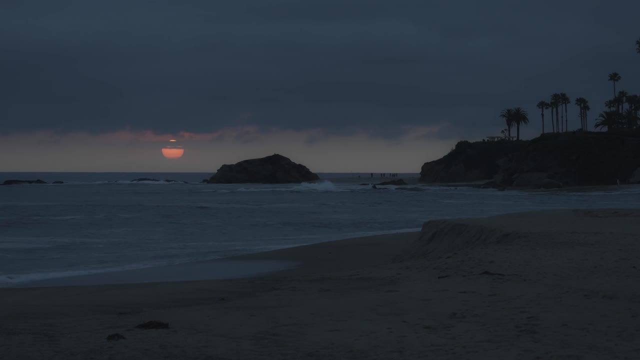 Timelapse of the sun setting behind ocean cliffs and swaying palms, casting a golden glow over the beach as evening falls on a tranquil coastal scene.