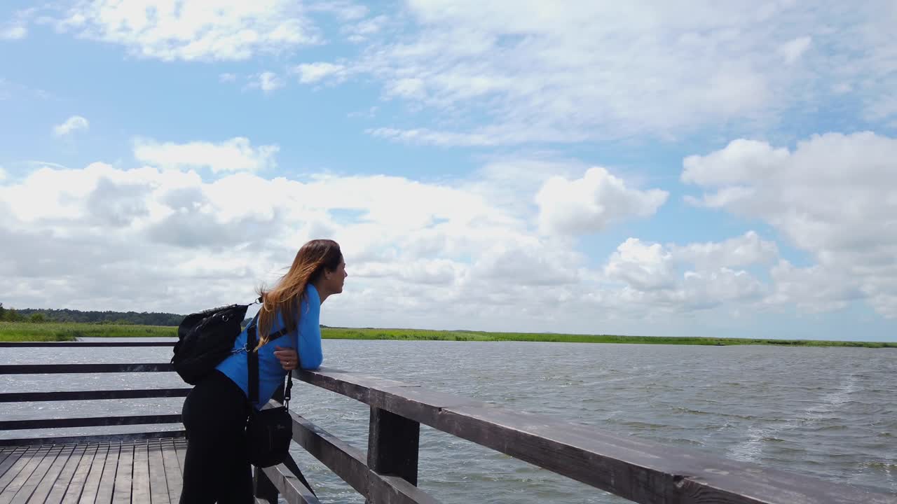 Female tourist with backpack admiring lake view from wooden pier, embracing tranquility of nature in Poland