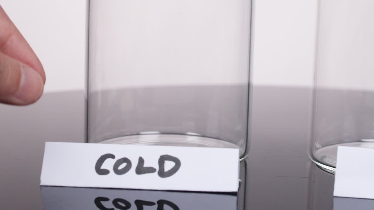 Hand places 'cold' label next to empty glass beaker on reflective laboratory surface, static camera
