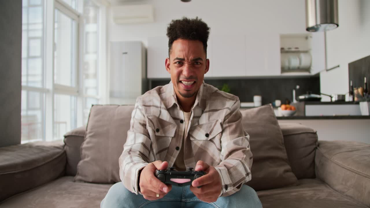 um jovem triste e zangado com pele preta uma morena com caule em uma camisa a quadras de cor creme senta-se em um sofá em um apartamento moderno e joga um jogo usando um joystick preto e está triste por sua derrota em um apartamento moderna