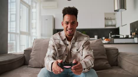 A-sad-and-angry-young-man-with-Black-skin-a-brunette-with-stubble-in-a-cream-colored-checkered-shirt-sits-on-a-sofa-in-a-modern-apartment-and-plays-a-game-using-a-black-joystick-and-is-sad-about-his-defeat-in-a-modern-apartment