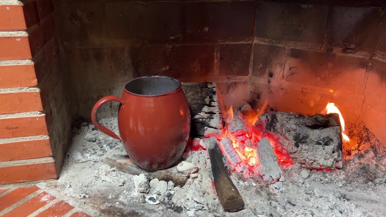 En person rører i indholdet af et rødt krus placeret over glødende kul i en muret pejs. Den rustikke ramme er ideel til temaer om varme og hygge.