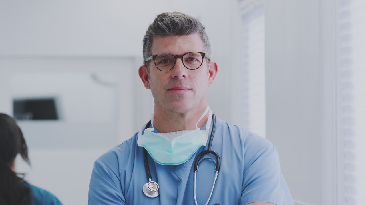 retrato de un médico masculino maduro vestido con trajes de exfoliación de pie en un concurrido pasillo del hospital