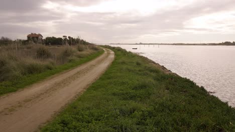 4K-aerial-view-of-a-dirty-road-in-the-bedside-of-Ria-de-Aveiro-in-the-estuary-of-river-Vouga,-drone-moving-forward,-boat-silhouette,-60fps