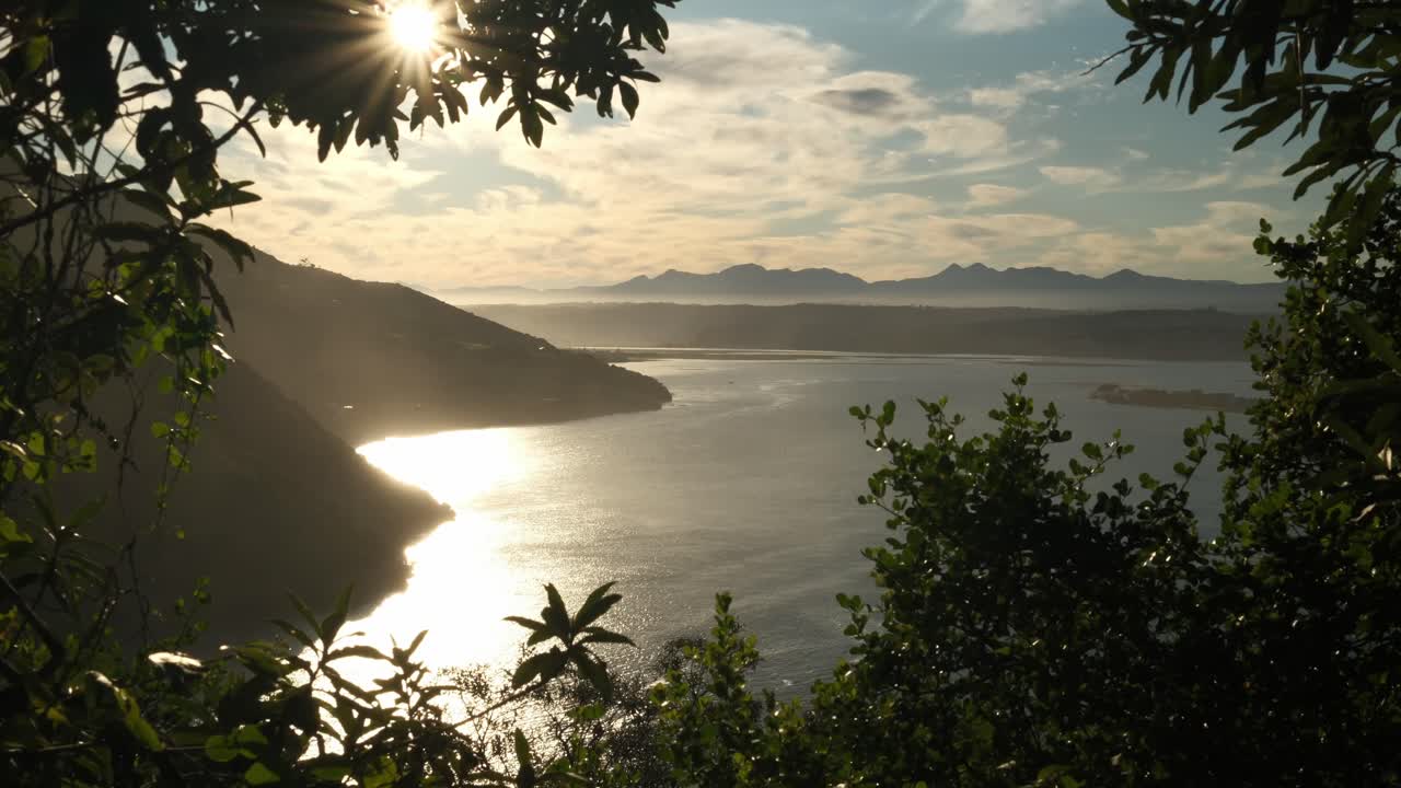 A perfectly framed static 4k shot of a beautiful peaceful lake. It is framed with tree branches. A hill and mountains are covered in golden hour light during a warm winters day in South Africa