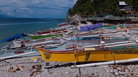 A-collection-of-many-kayak-style-fishing-boats-on-a-sandy-beach-in-Mabua,-Philippines-with-turquoise-water-and-lush-jungle-in-the-background