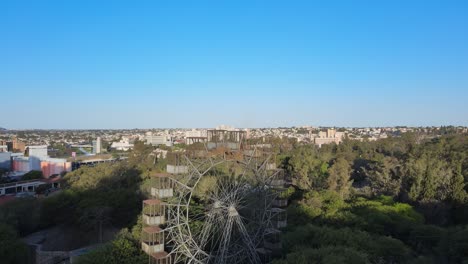 drone reverse flying overlooking distance downtown cityscape, reveals iconic landmark rusty old ferris wheel, the creation of gustave eiffel in famous urban sarmiento park in cordoba, argentina