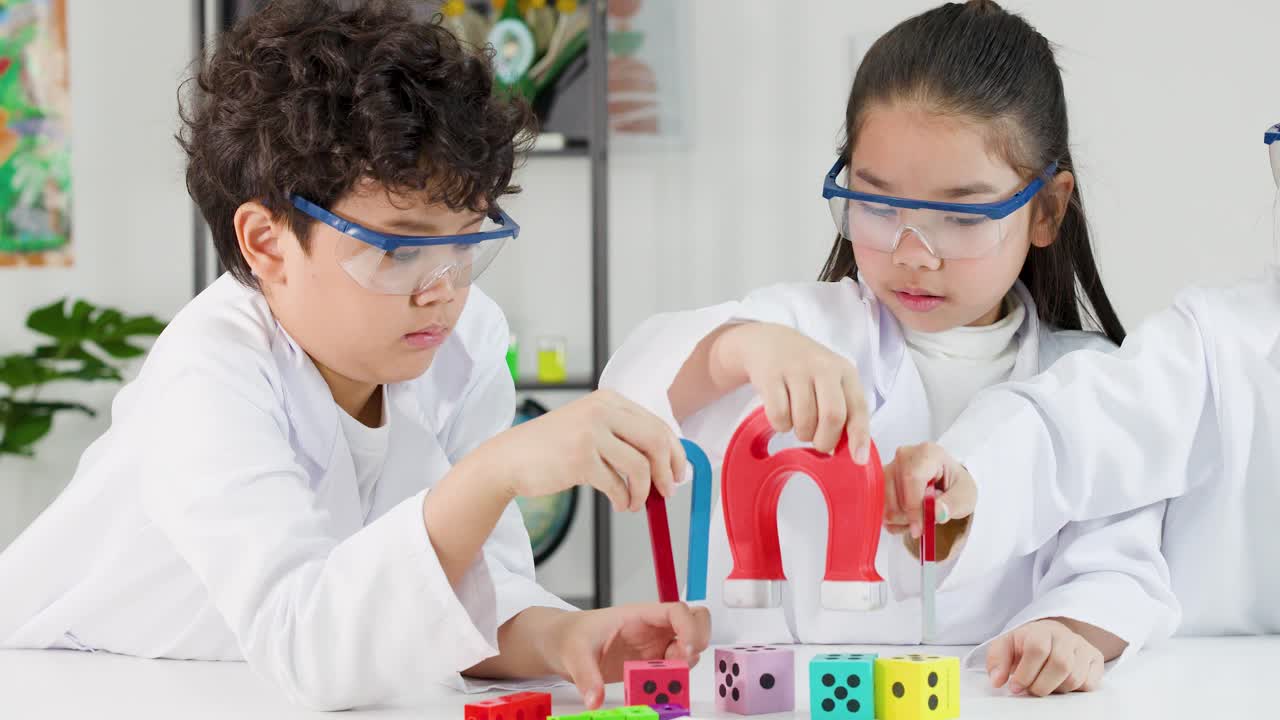 Dua siswa sekolah dasar mengenakan kacamata pelindung dan jas lab saat menjelajahi magnetisme dengan magnet tapal kuda.