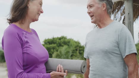 happy senior caucasian couple holding yoga mat and talking on beach, in slow motion