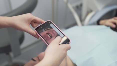dentist in modern dental clinic uses her mobile phone camera to take pictures of teeth of her male patient after successful treatment