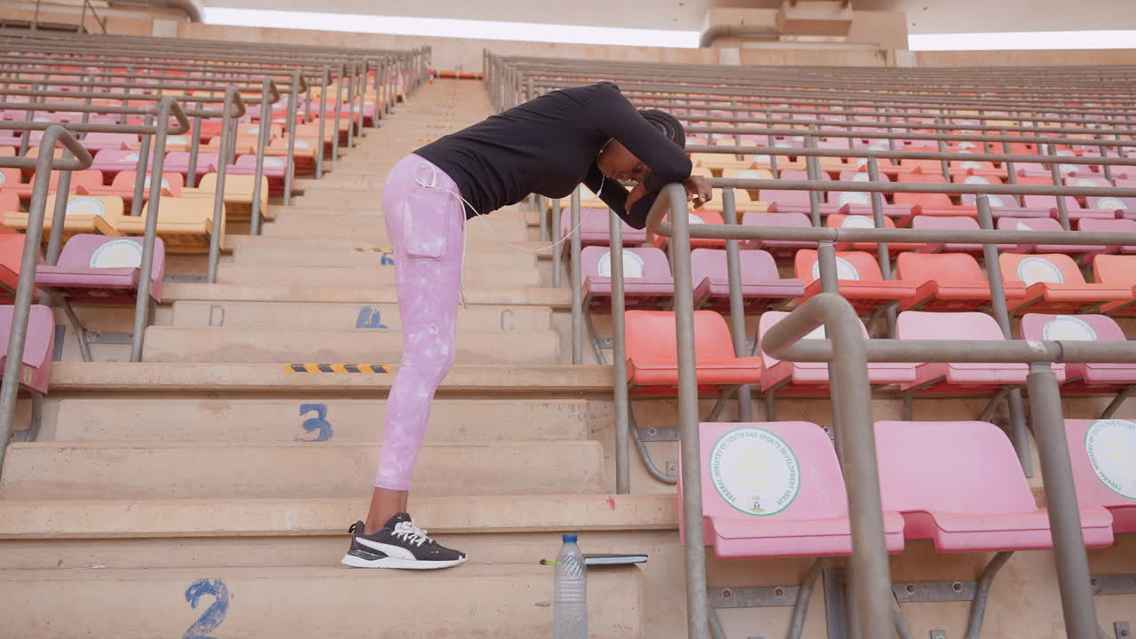 tired woman leaning on stadium railing, resting after intense training session on vacant bleachers, pink pants and sneakers, concrete steps and metal railing, heavy breath and paused posture conveying