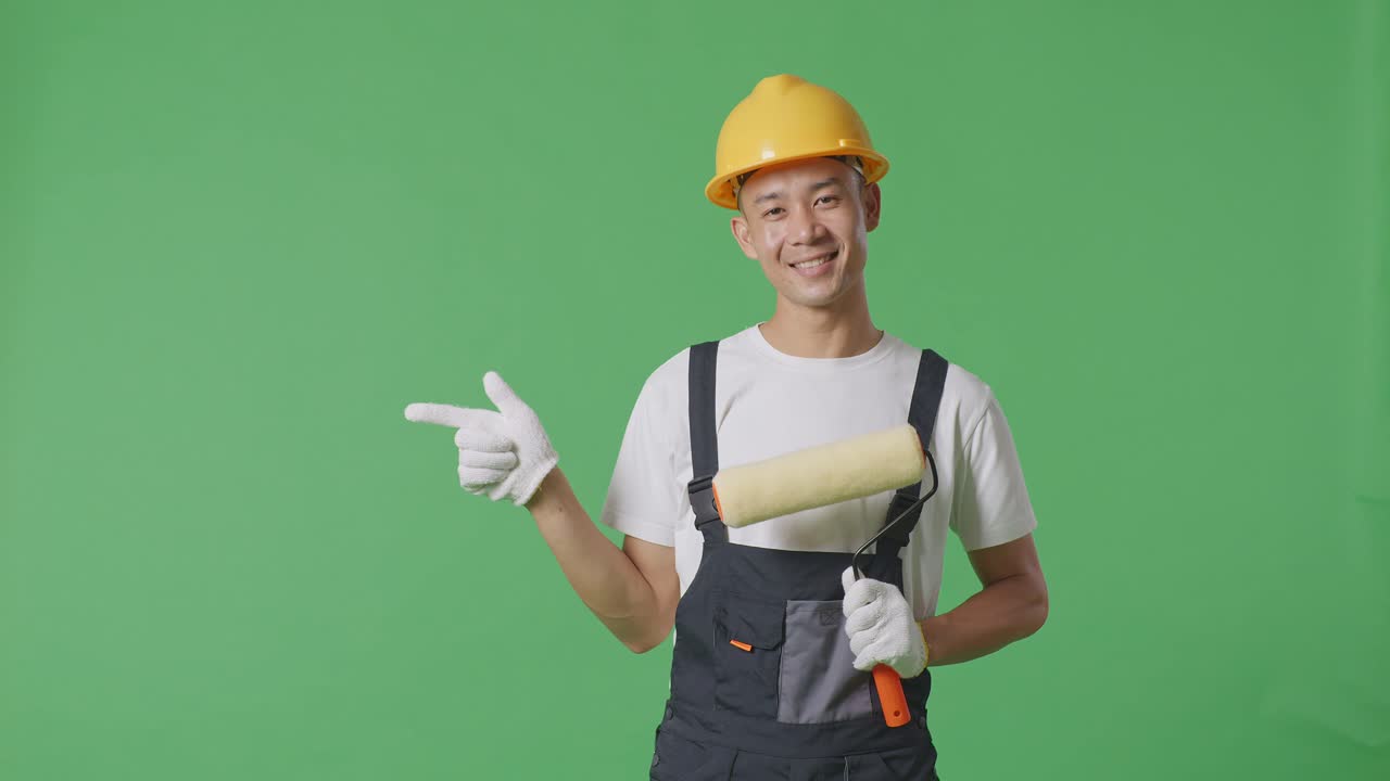Asian Man Painter Wearing Safety Helmet Smiling And Pointing To Side While Standing In The Green Screen Background Studio