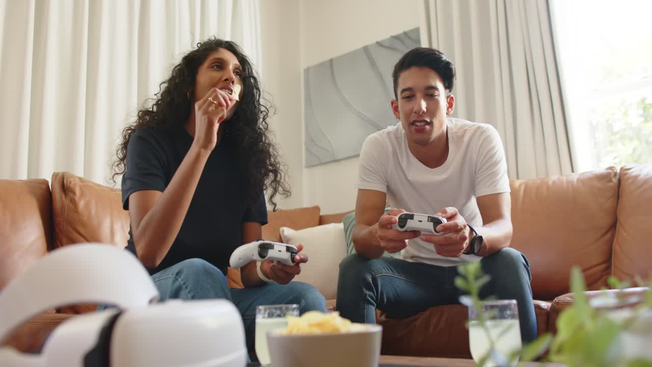 Couple starting console game, playing with white controllers on couch, sharing chips, relaxing